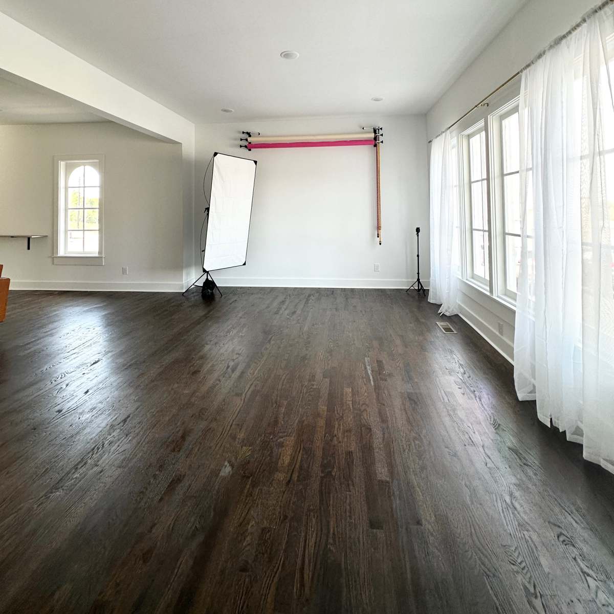 A spacious, empty room with dark hardwood floors and large windows covered by sheer white curtains, featuring a backdrop setup on one wall.