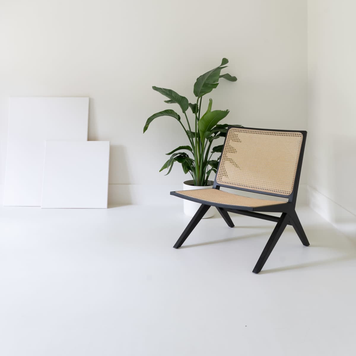 A minimalist room features a black-framed chair with a woven seat, accompanied by a potted plant and stacked white canvases against a light-colored wall.