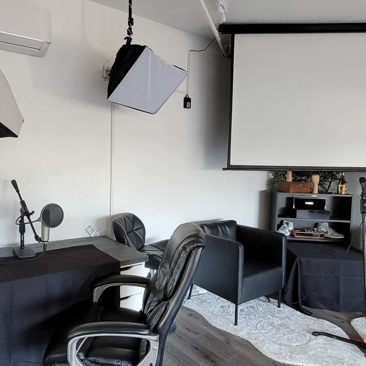 A studio space with a desk, microphone, camera equipment, two chairs, and a projector screen mounted on the wall.