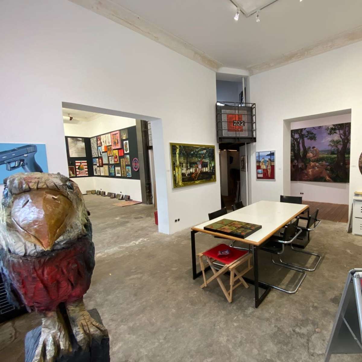 An art gallery interior with a central table surrounded by chairs, featuring various artworks displayed on the walls.