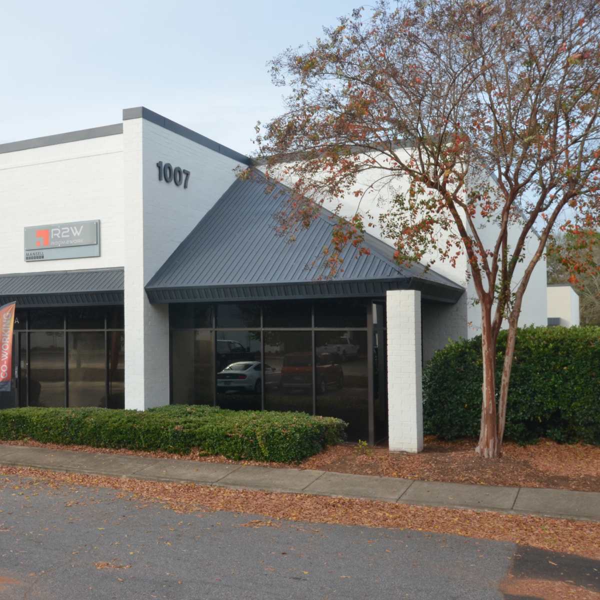 A white commercial building with a dark gray metal roof, featuring large windows and a sign that reads "R2W," located on a clean street.