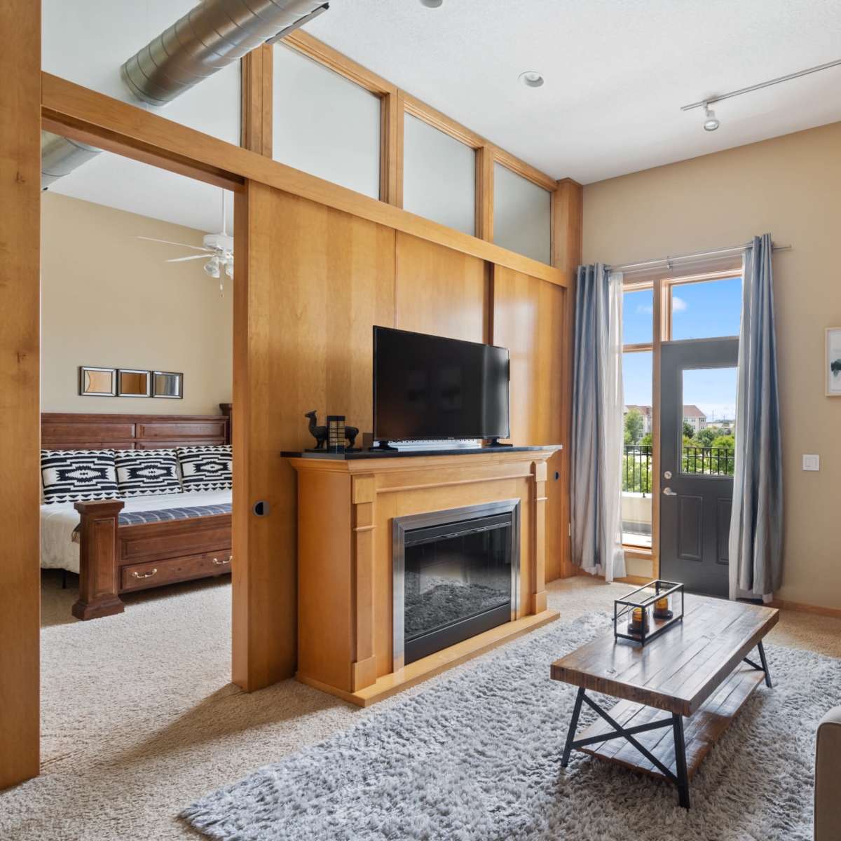 A cozy living room with a television mounted on a wooden mantel, a gray sofa, and large windows that provide natural light.