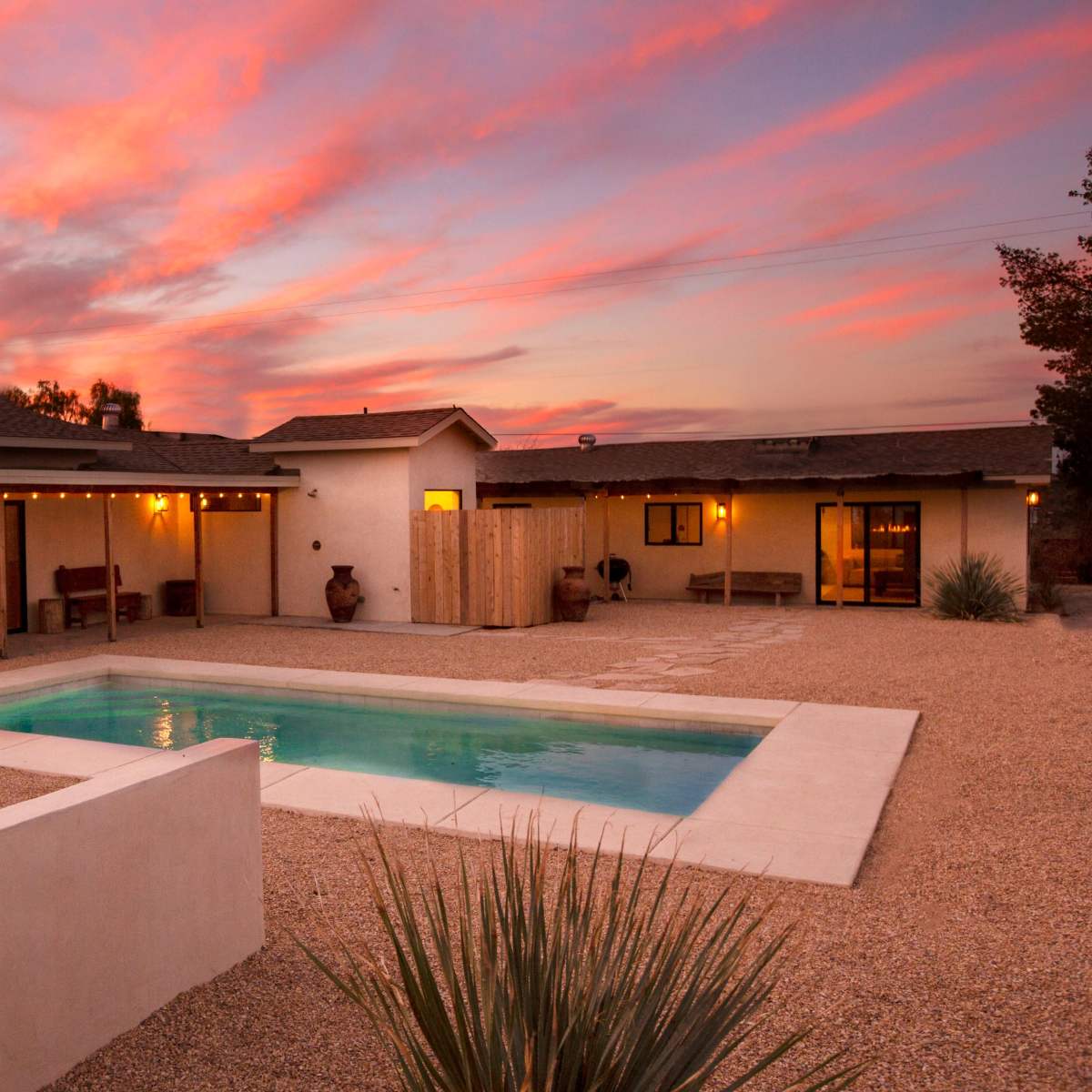A modern home with a pool and fire feature is surrounded by a gravel courtyard under a sunset sky.