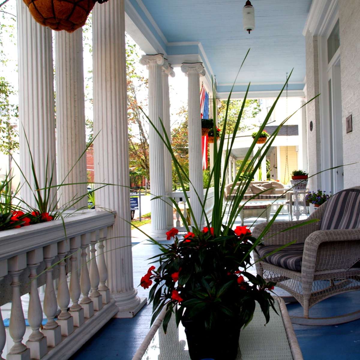 A porch with white columns, potted plants, and a seating area featuring wicker furniture and a glass table.