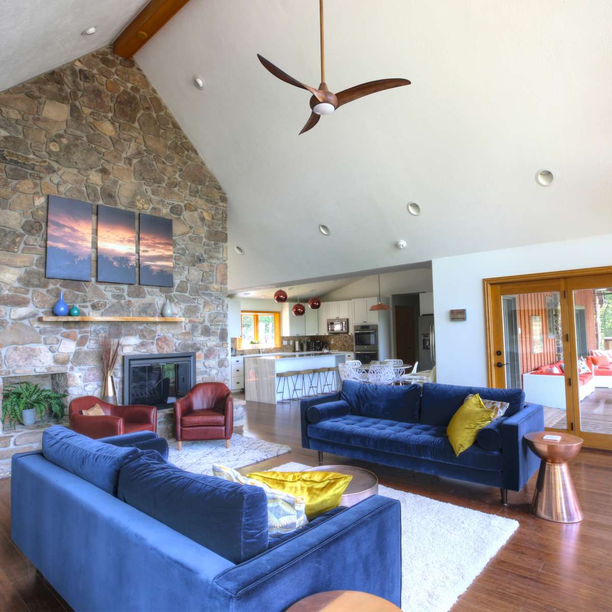 A spacious living room featuring a stone fireplace, two blue sofas, and a dining area in the background with large windows.