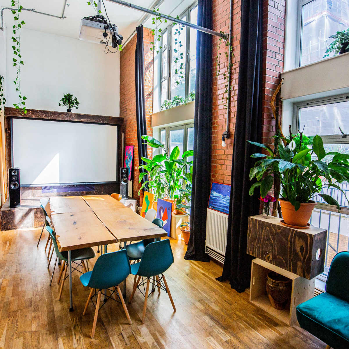 A well-lit meeting room features a wooden table surrounded by blue chairs, with large windows, green plants, and a projector screen at the front.