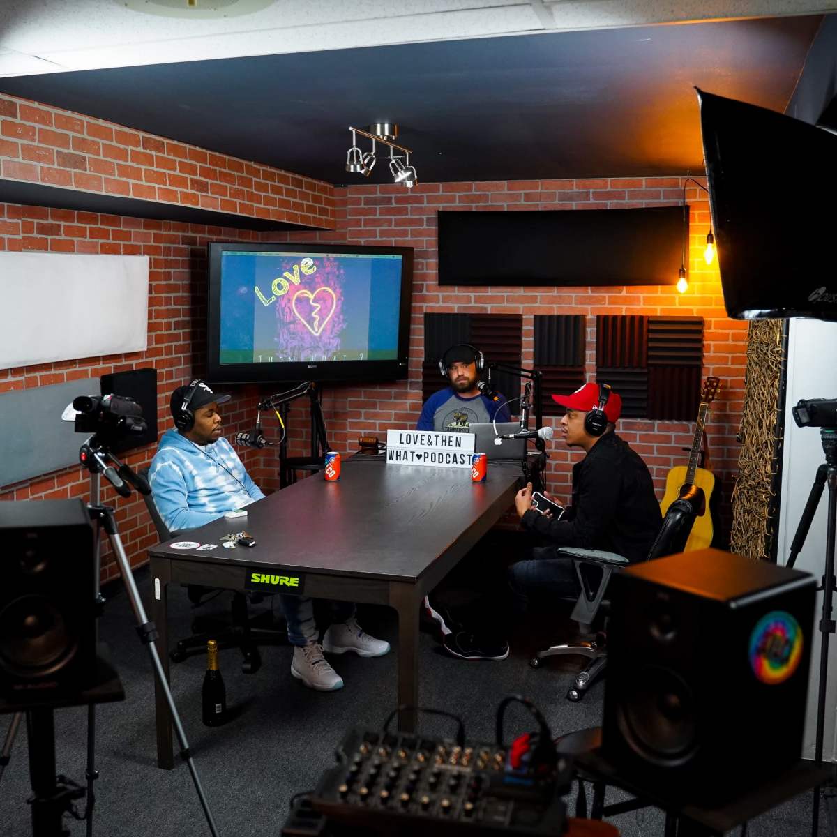 Three individuals are seated at a table in a podcast studio, equipped with microphones and cameras, discussing in front of a monitor displaying a colorful heart graphic.