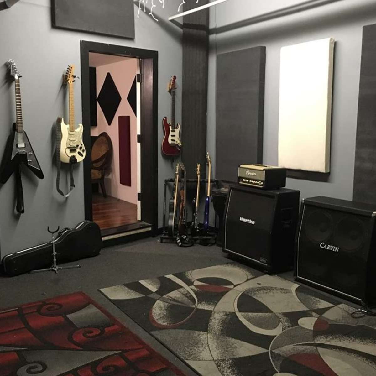 A music rehearsal room with electric guitars mounted on the wall, amplifiers positioned on the floor, and a patterned area rug.