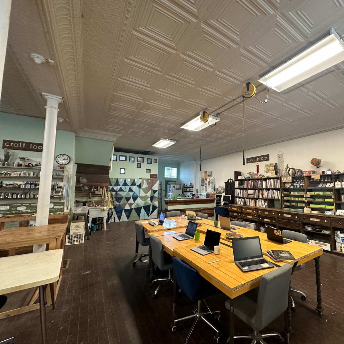 A spacious workshop with a long wooden table surrounded by laptops, bookshelves filled with materials, and various crafting supplies along the walls.