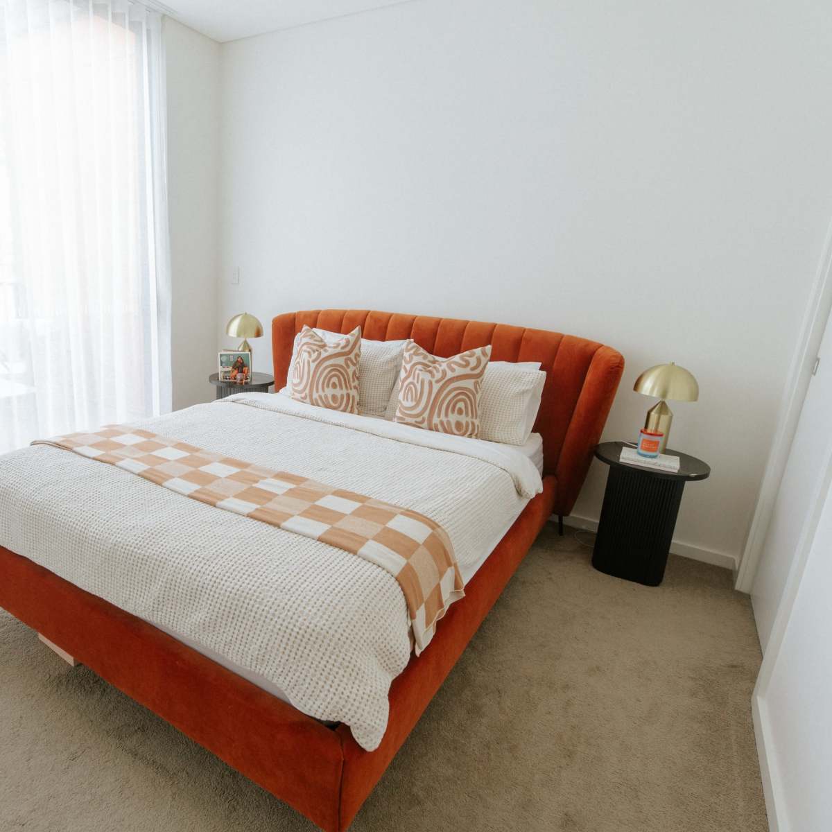The bedroom features an orange upholstered bed with layered bedding, two patterned pillows, and a bedside table with a lamp.