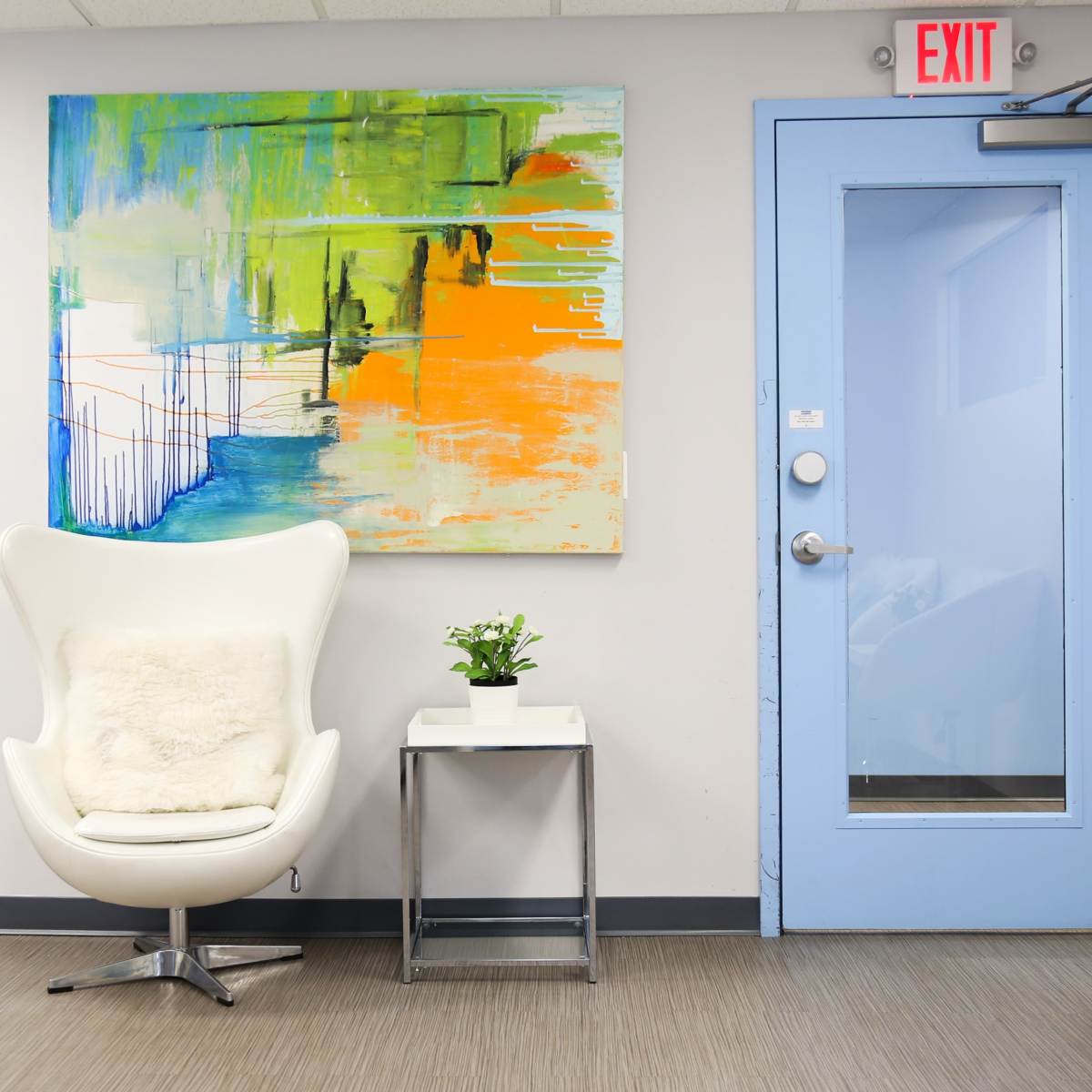 A modern waiting area featuring a colorful abstract painting, a white chair with a fur throw, a small plant on a glass table, and a blue door leading to another room.