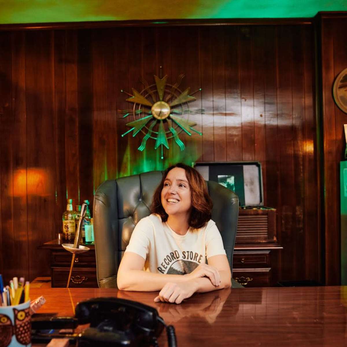 A woman sits at a wooden desk in a warmly lit office with vintage decor, smiling and looking towards the camera.