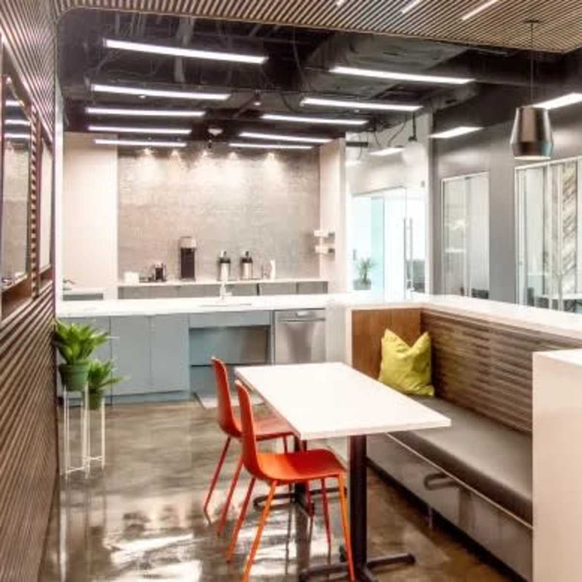 A modern office kitchen area featuring a dining table with red chairs, a bench seat, and a light-colored kitchen cabinetry and appliances.
