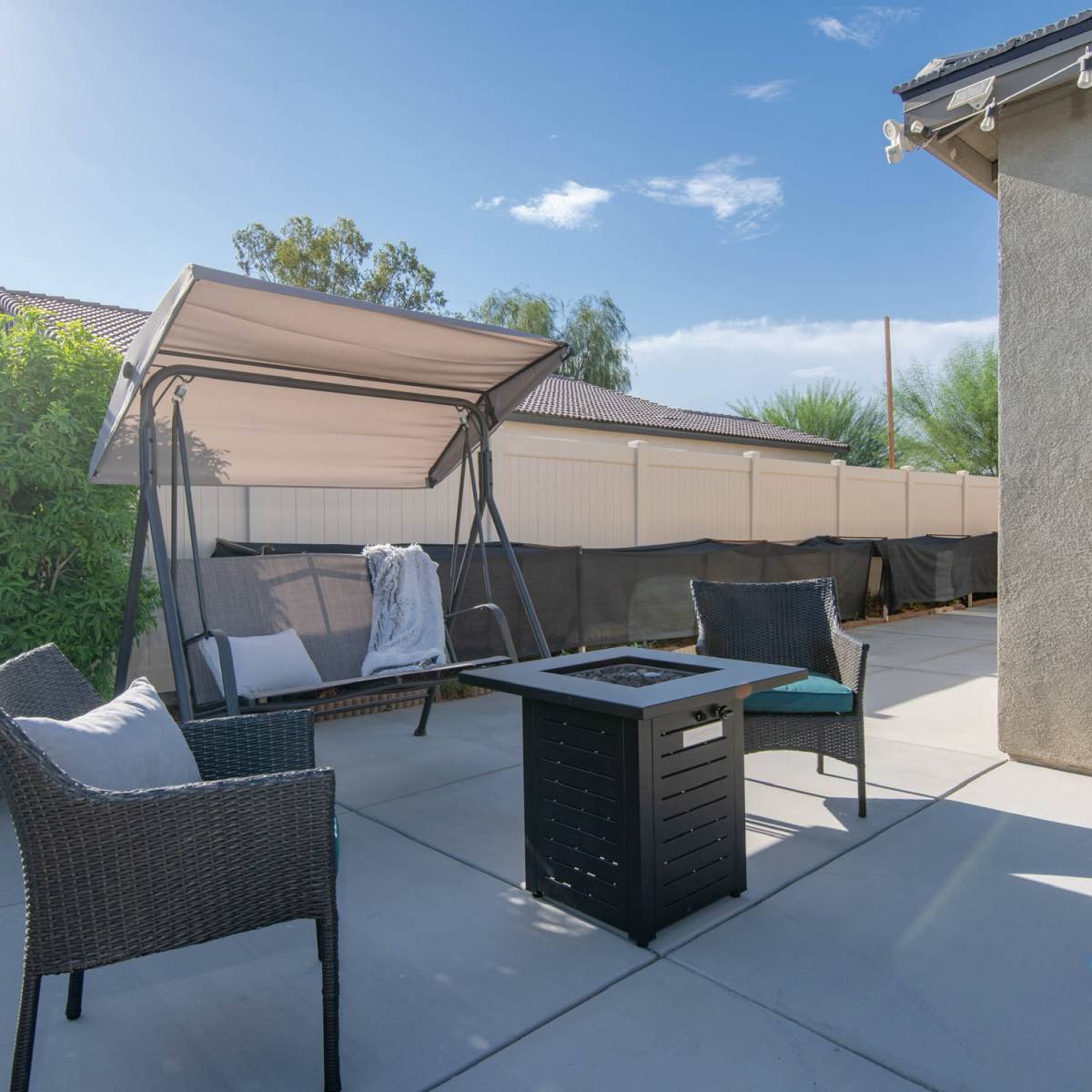 A covered swing and two chairs surround a rectangular fire pit on a concrete patio in a backyard.