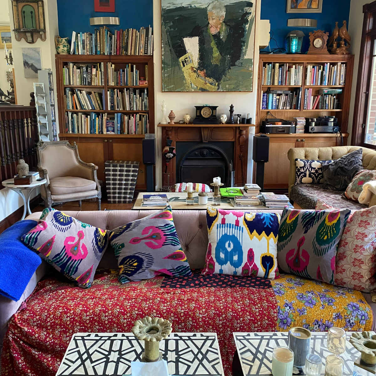 A cozy living room with a mix of bookshelves, colorful cushions on sofas, a decorative fireplace, and a variety of artworks on the walls.