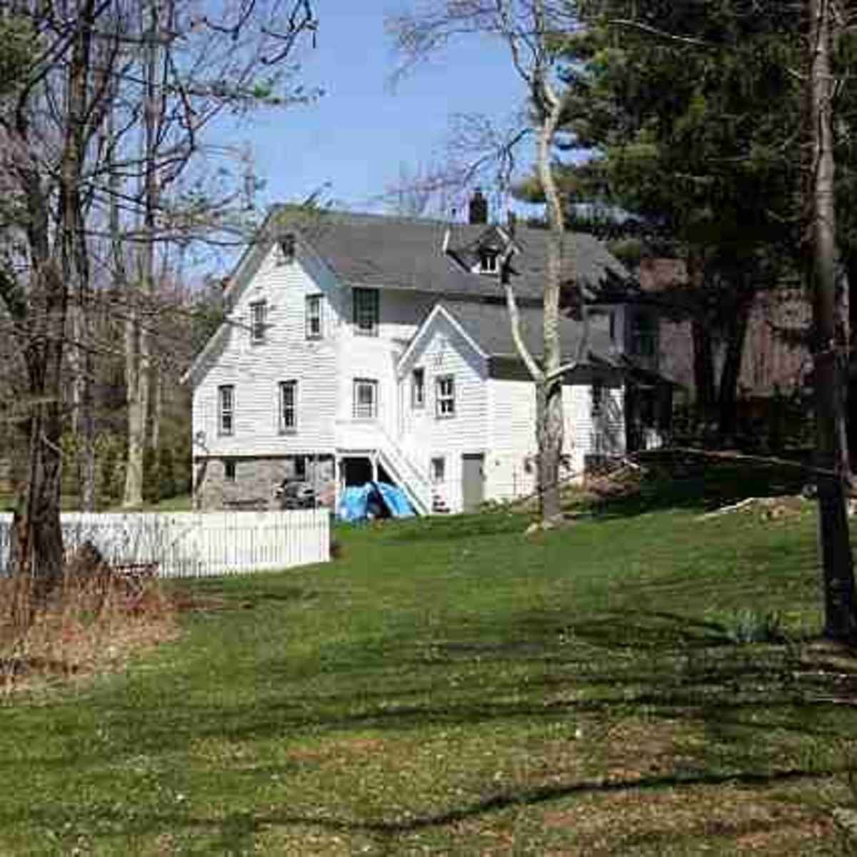 A large, two-story house is situated near a white picket fence, surrounded by trees and green grass on a sunny day.