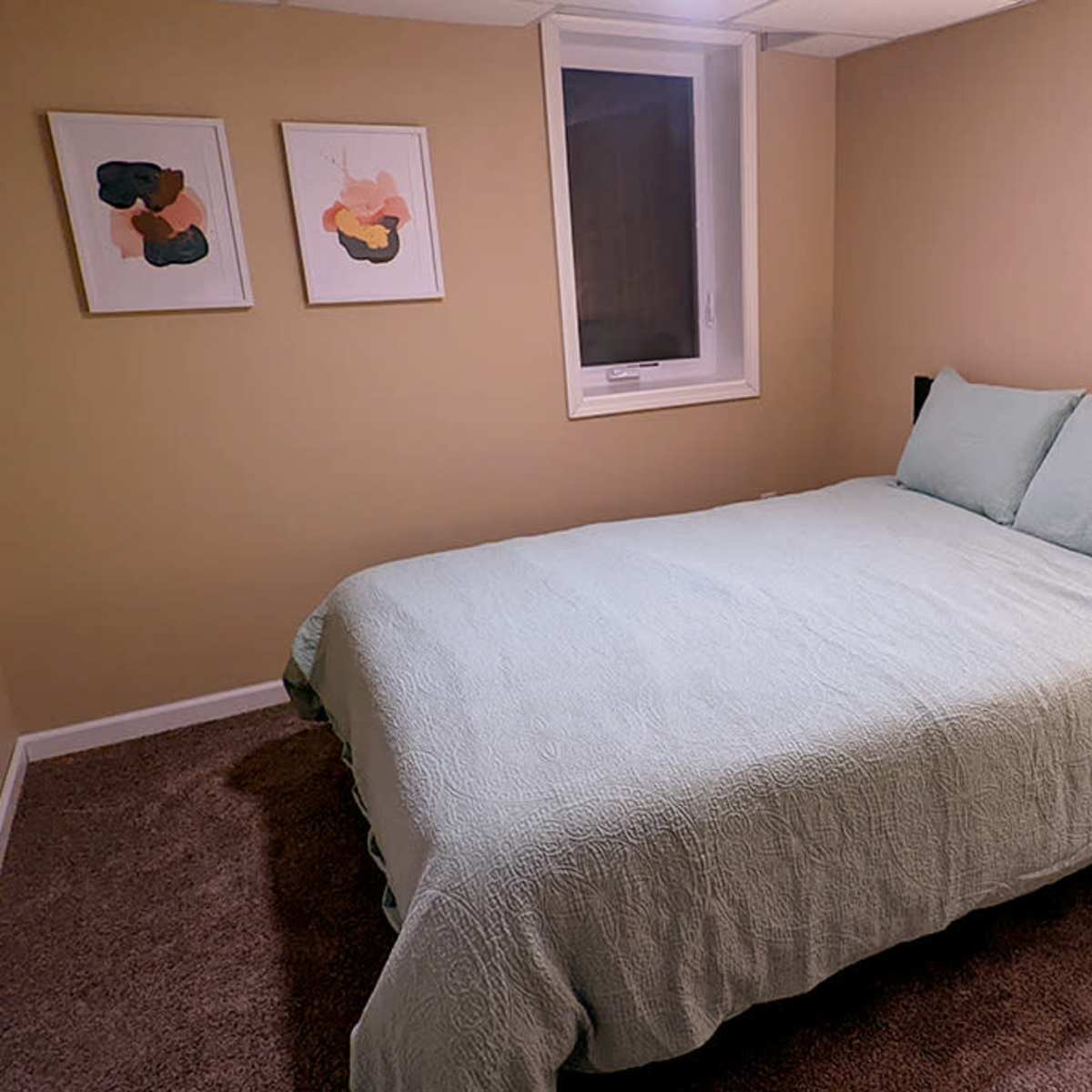 A small bedroom with a light green bedspread, a bedside table with a lamp, and two abstract art pieces hanging on the wall.