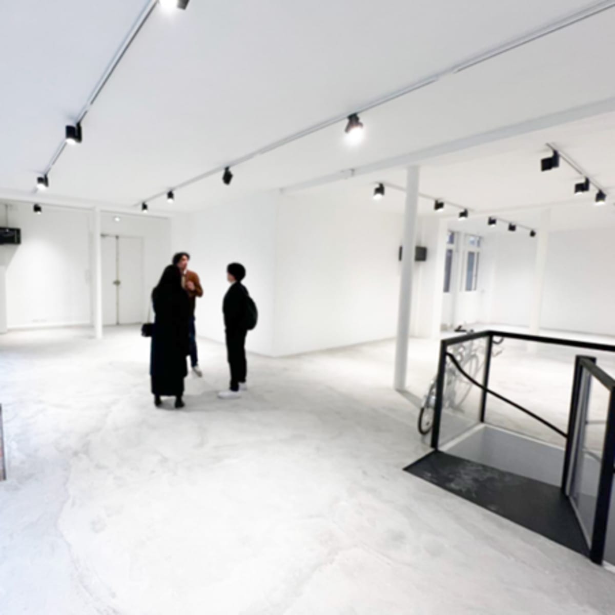 Three people stand conversing in an empty, brightly lit gallery space with white walls and a concrete floor.