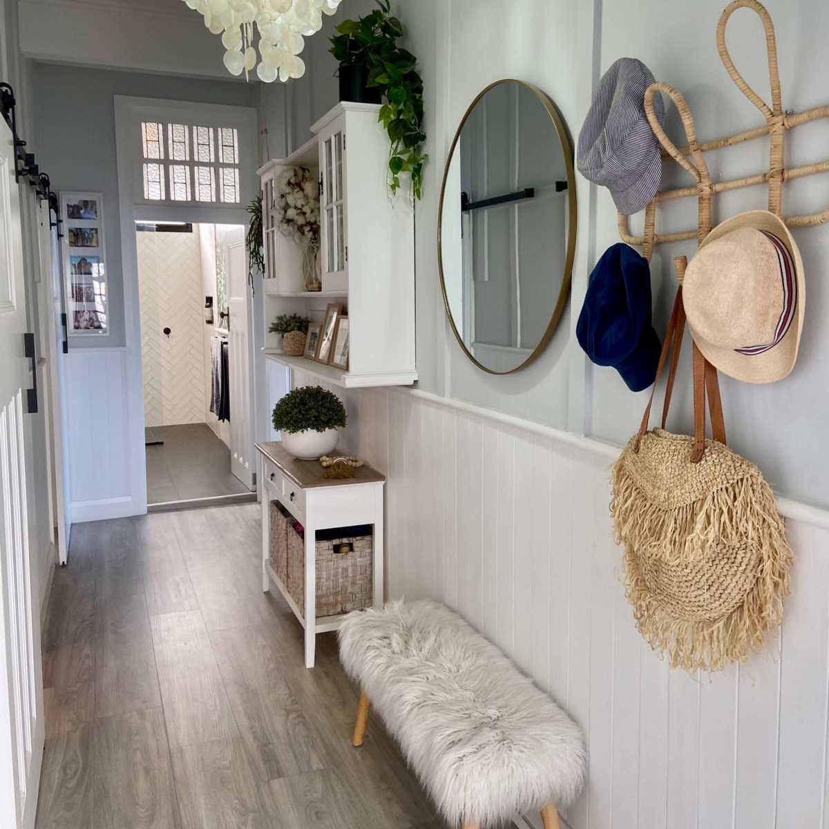 A narrow hallway featuring a white bench with a fluffy cushion, a round mirror on the wall, various hats hanging on hooks, and a light fixture above.