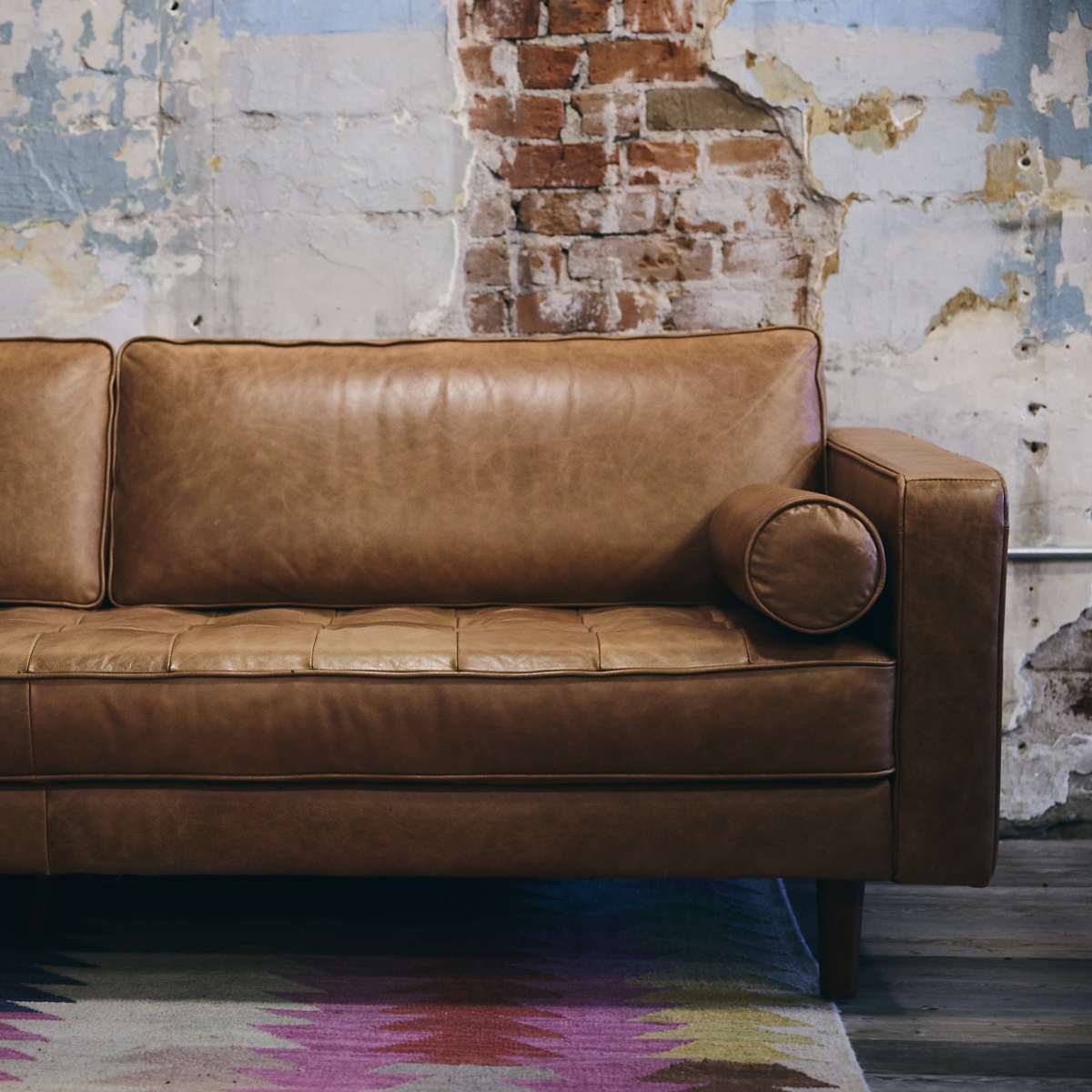 A brown leather sofa with a round armrest is positioned against a wall featuring peeling paint and exposed brick.