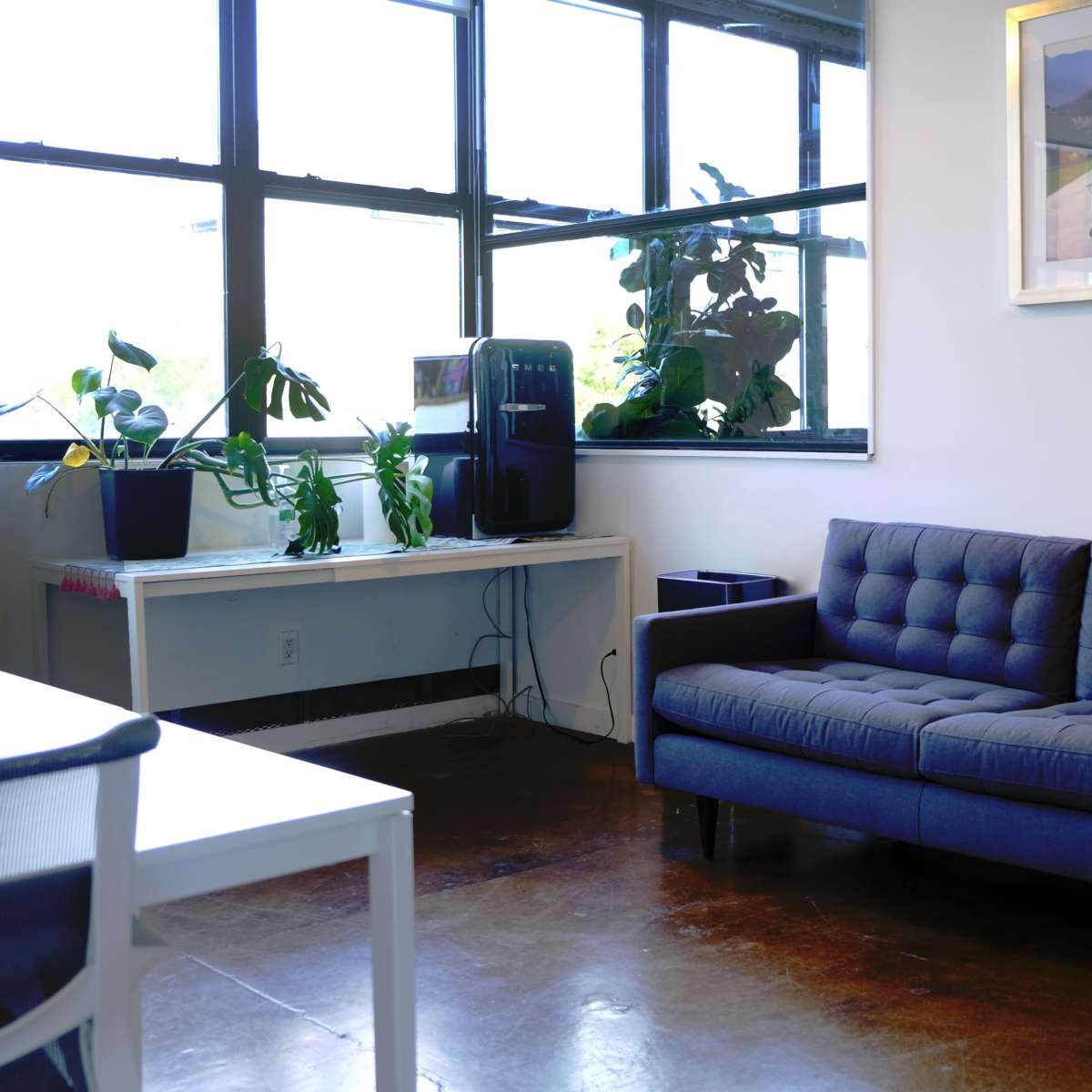 A modern living room with a blue couch, a white table, and large windows allowing natural light, alongside potted plants and a water cooler.