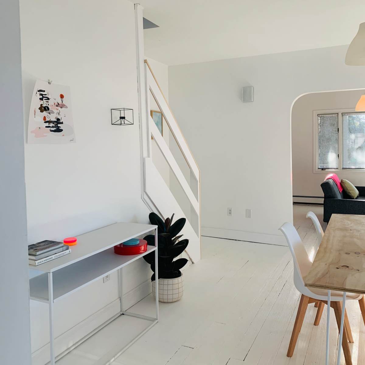A bright, minimalist interior of a room featuring a white wall and wooden staircase, with a simple table and chairs set up near a plant.
