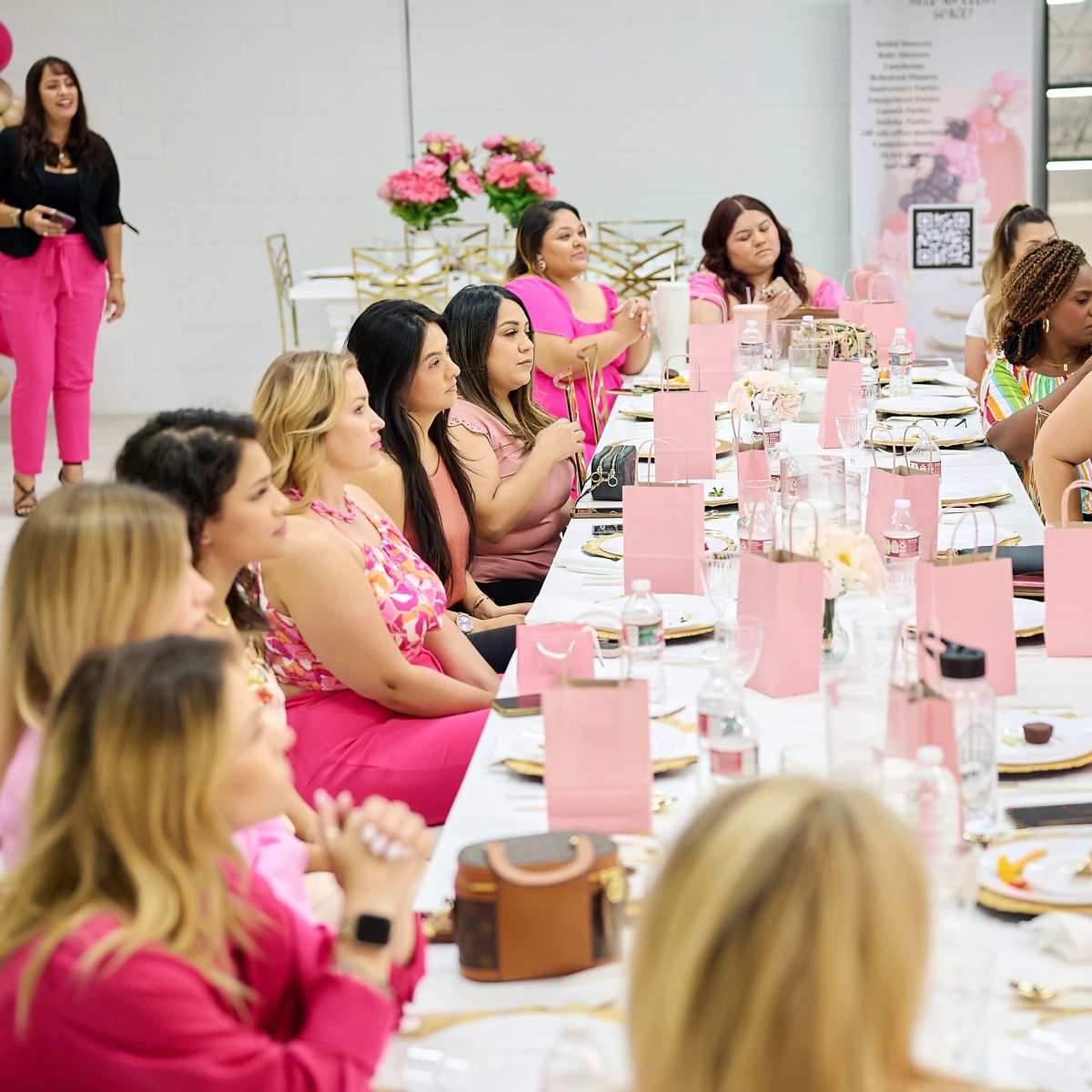 A group of women in pink attire are seated at a long table adorned with pink decorations, attentively watching a speaker at a festive event.