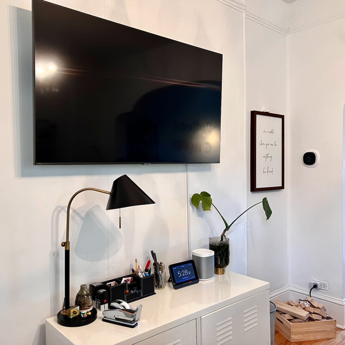 A mounted flat-screen TV above a white cabinet, with a desk lamp, various office supplies, a tablet, and a potted plant on the cabinet's surface.
