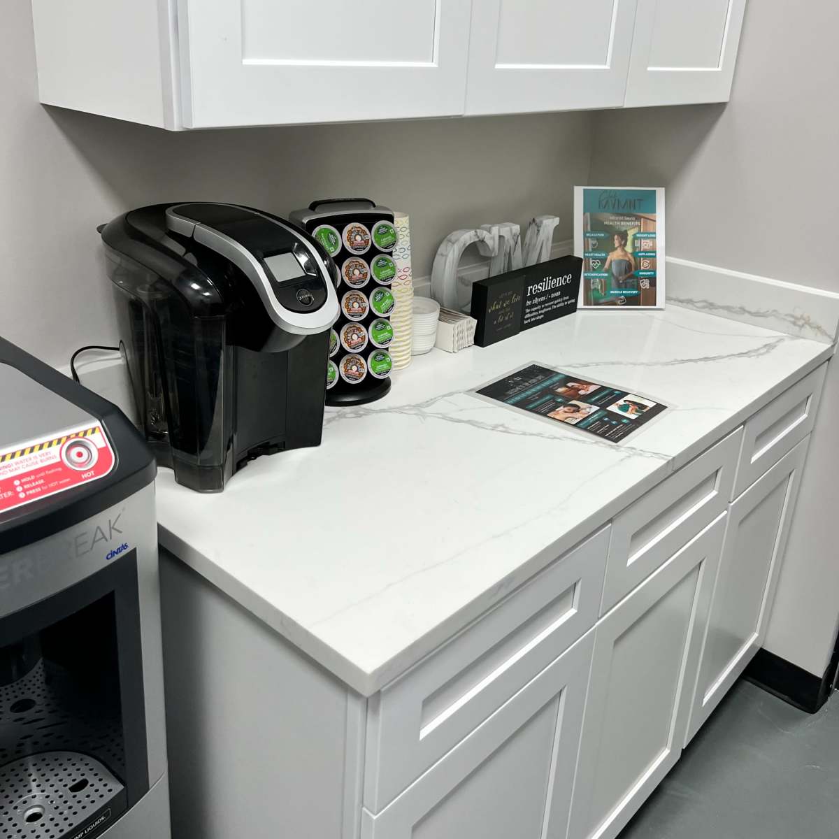 A clean countertop with a coffee maker, a carousel of coffee pods, a water pitcher, and a stack of brochures on a surface with a light-colored stone finish.