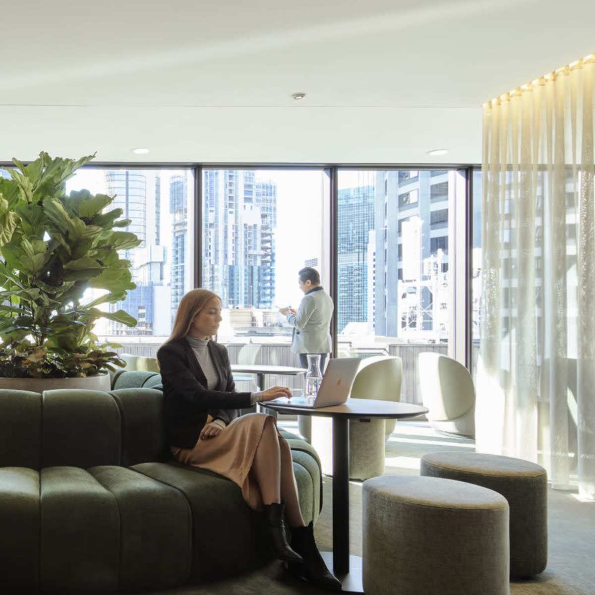 A modern workspace with a circular green seating area, large windows featuring a cityscape, and individuals working on laptops.