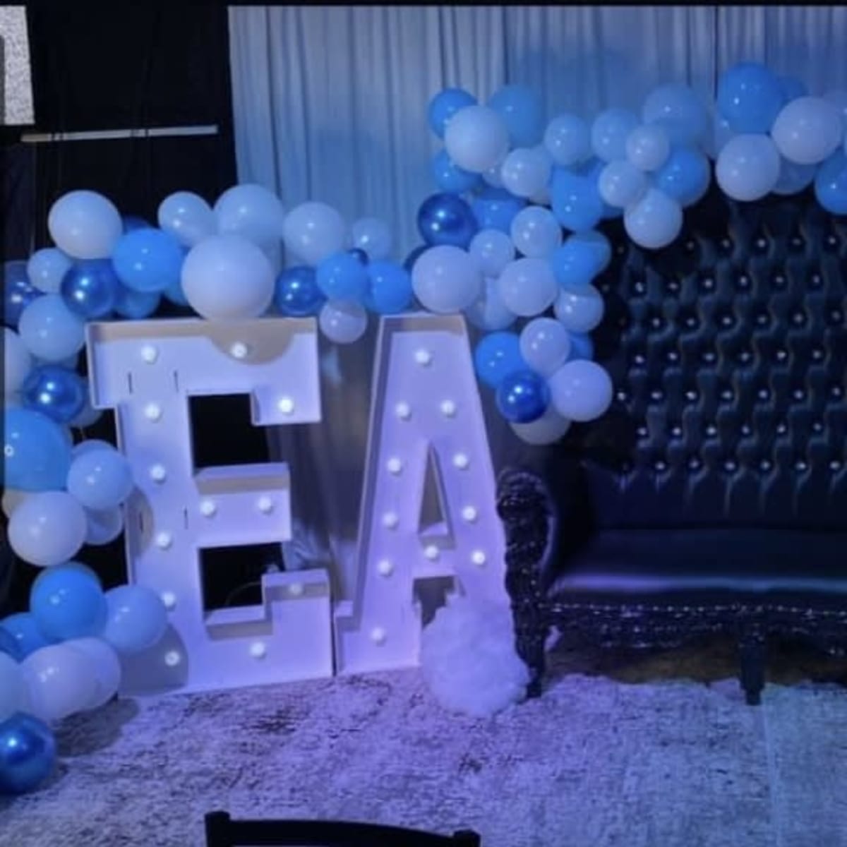 A black tufted sofa is surrounded by blue and white balloons, with illuminated letters "EA" displayed nearby.