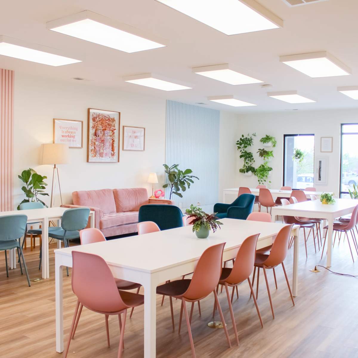 A bright and modern co-working space featuring white tables, colorful chairs, and large windows with plants decorating the interior.