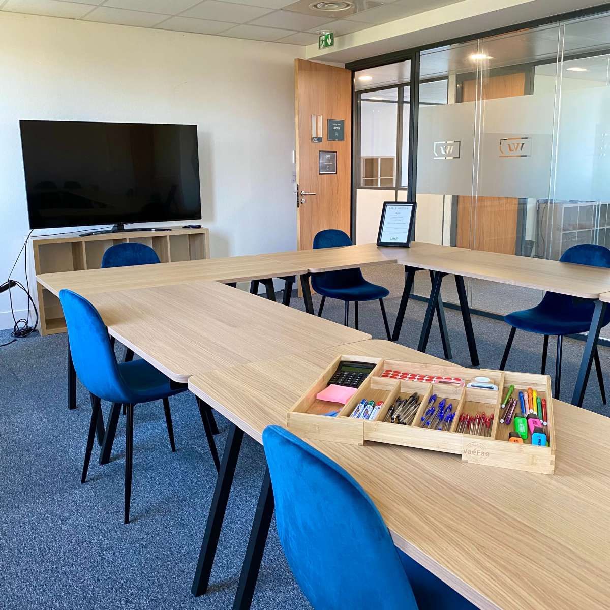 A modern conference room features a long wooden table surrounded by blue chairs, with a TV display and a tray of office supplies in the center.