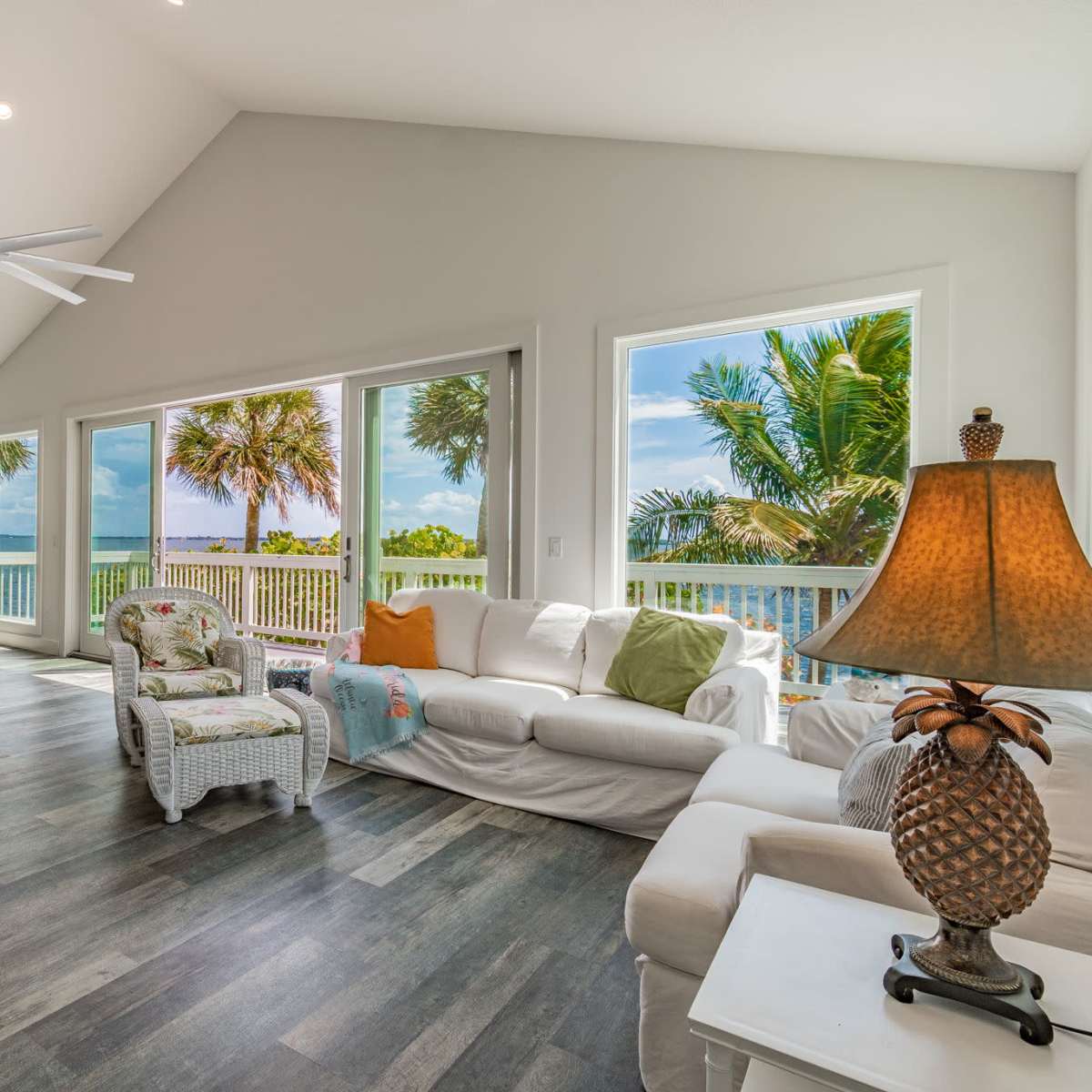 A spacious living room features large windows with views of palm trees and water, furnished with white sofas and a wicker chair.