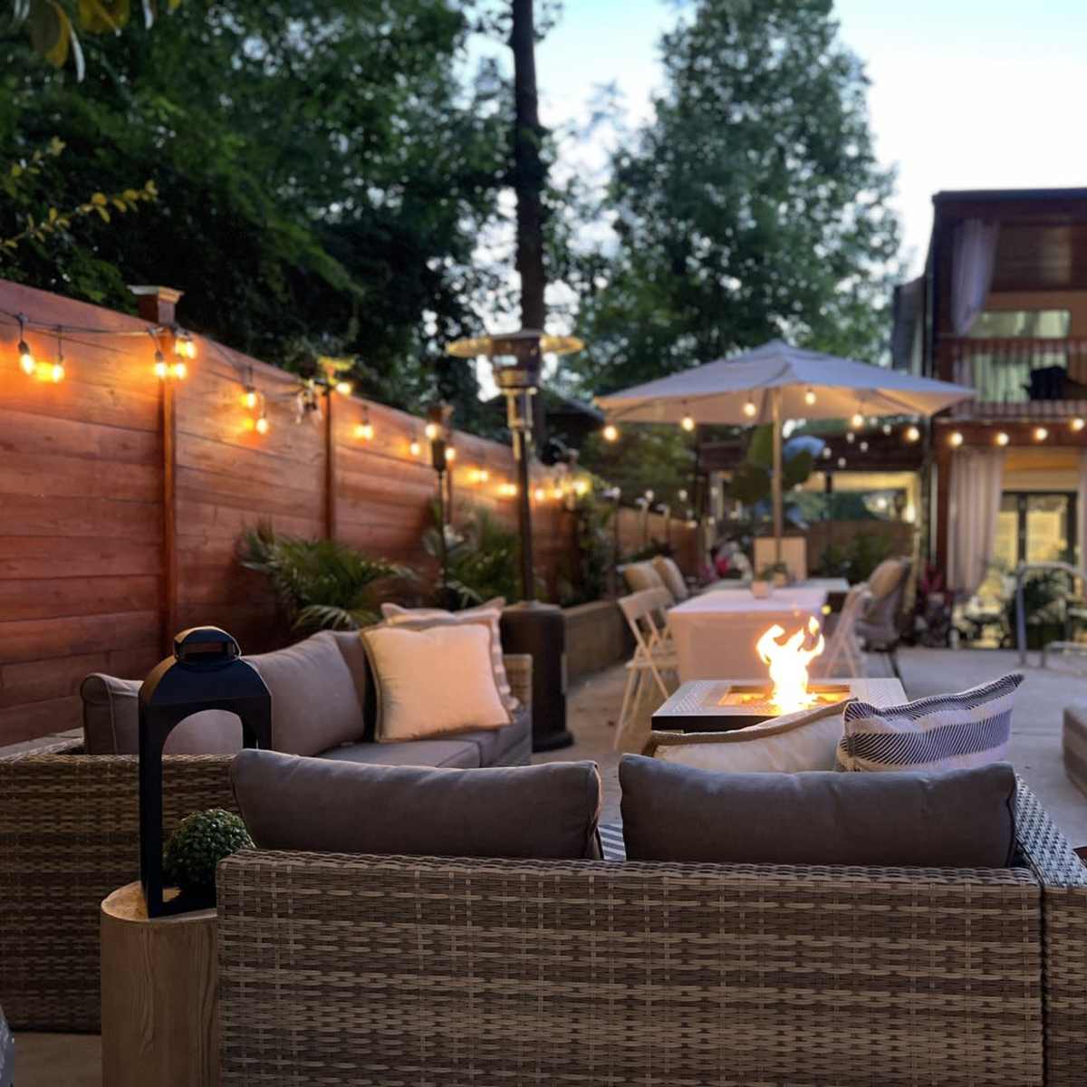 A cozy outdoor seating area with a fire pit, surrounded by a pool and string lights, in a backyard setting.