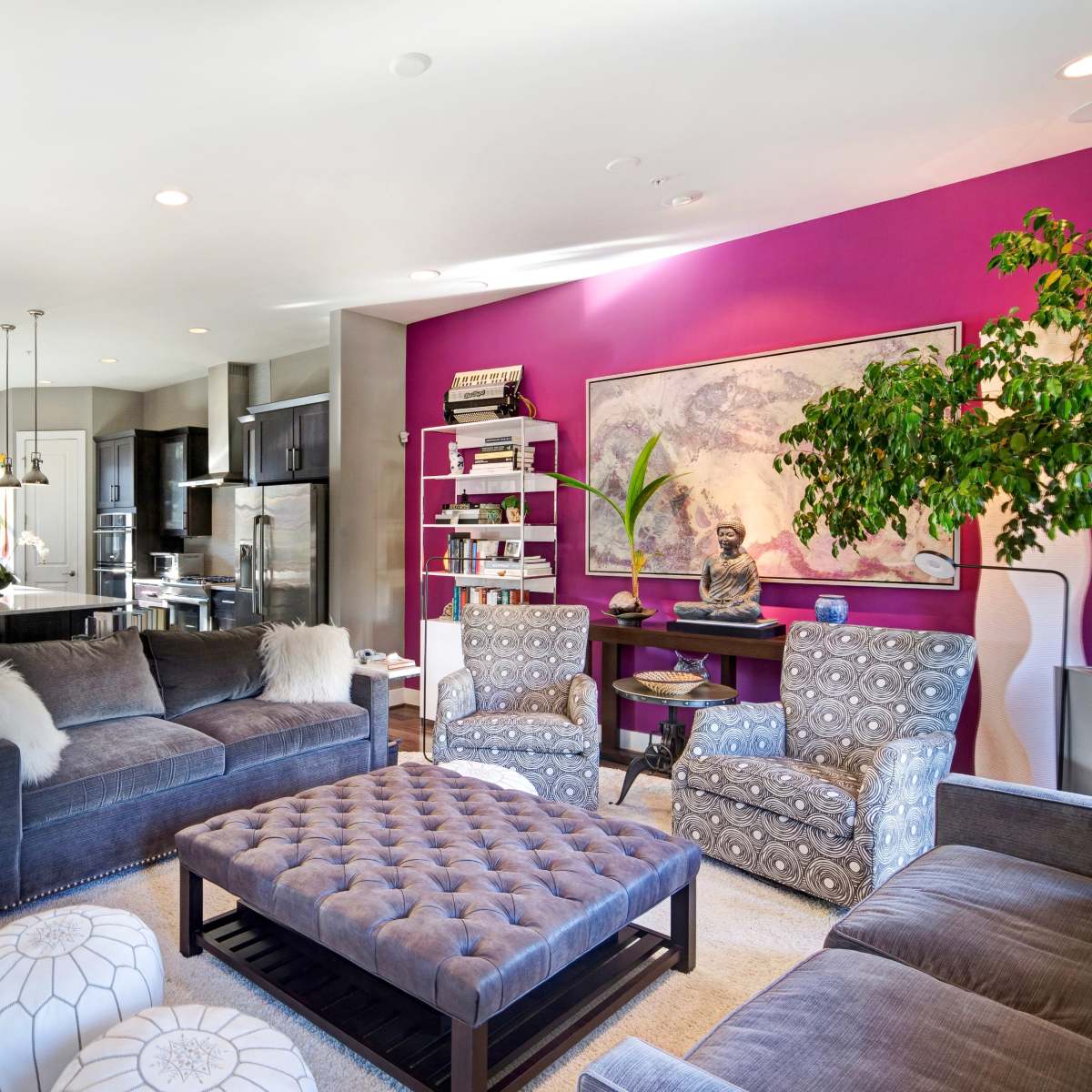 A contemporary living room featuring a plush gray sofa, patterned armchairs, a tufted coffee table, and bold purple accent walls, with a dining area in the background.