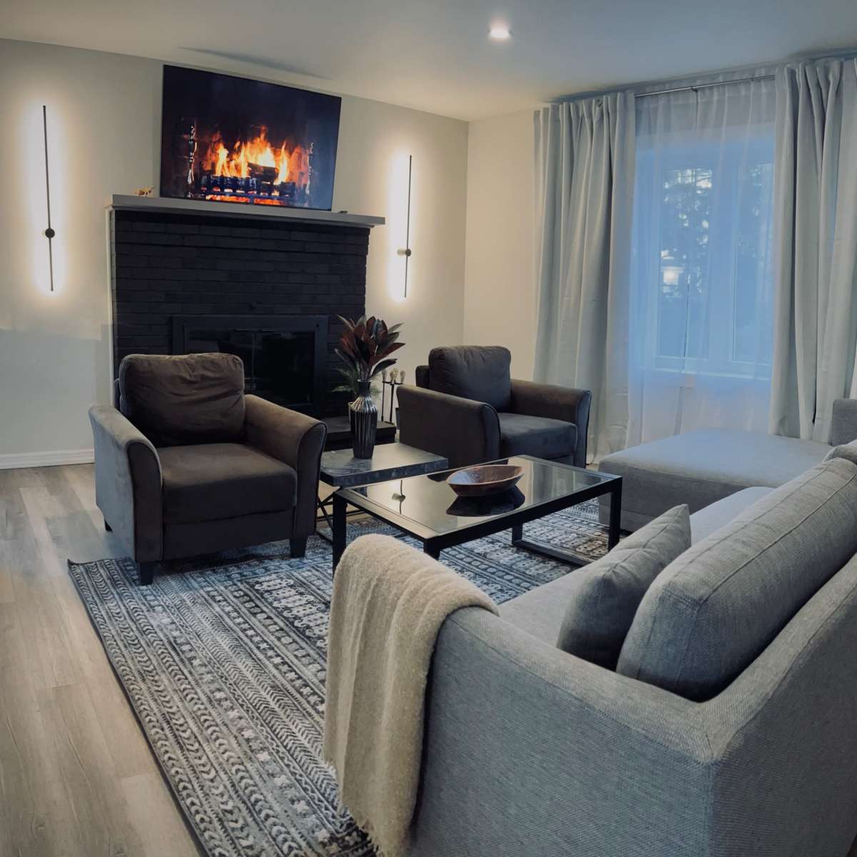 A cozy living room with a gray sofa, two armchairs, a coffee table, and a wall-mounted television displaying a fireplace.