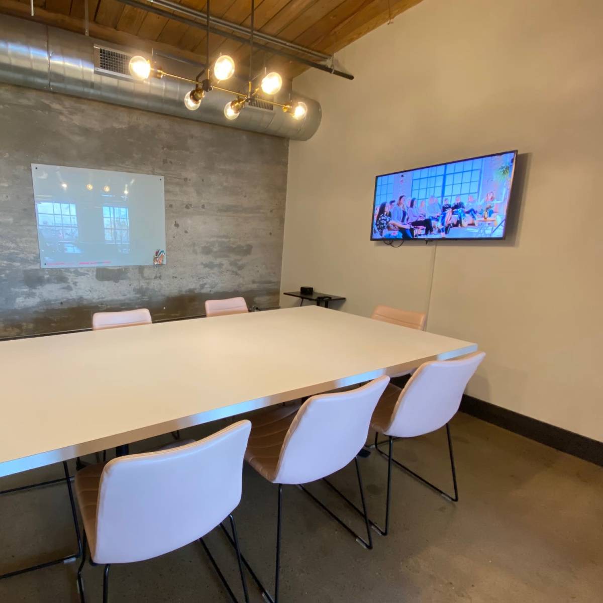 A modern conference room features a long white table surrounded by pink chairs, with a wall-mounted TV and a whiteboard.
