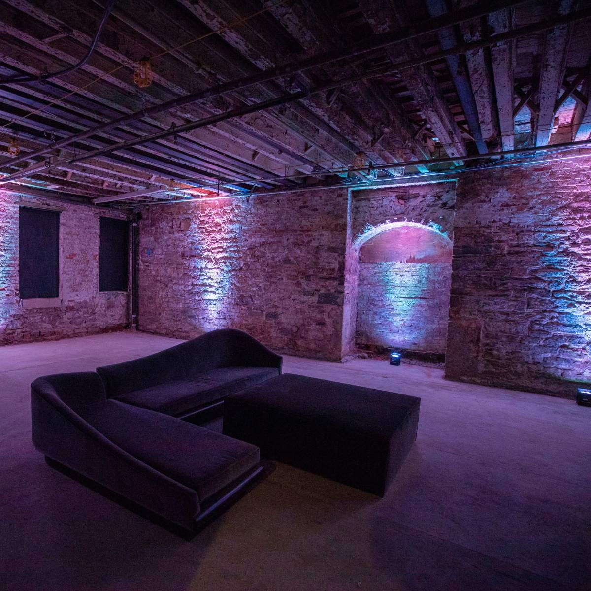 A dimly lit basement room with exposed brick walls and colorful lighting, featuring a modern black sofa set.