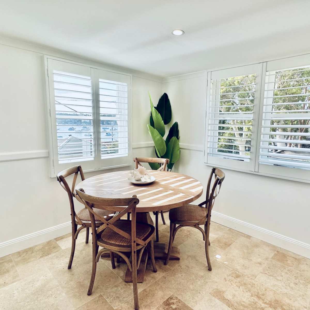 A round wooden dining table with four chairs is positioned near a window surrounded by white shutters and a large potted plant.