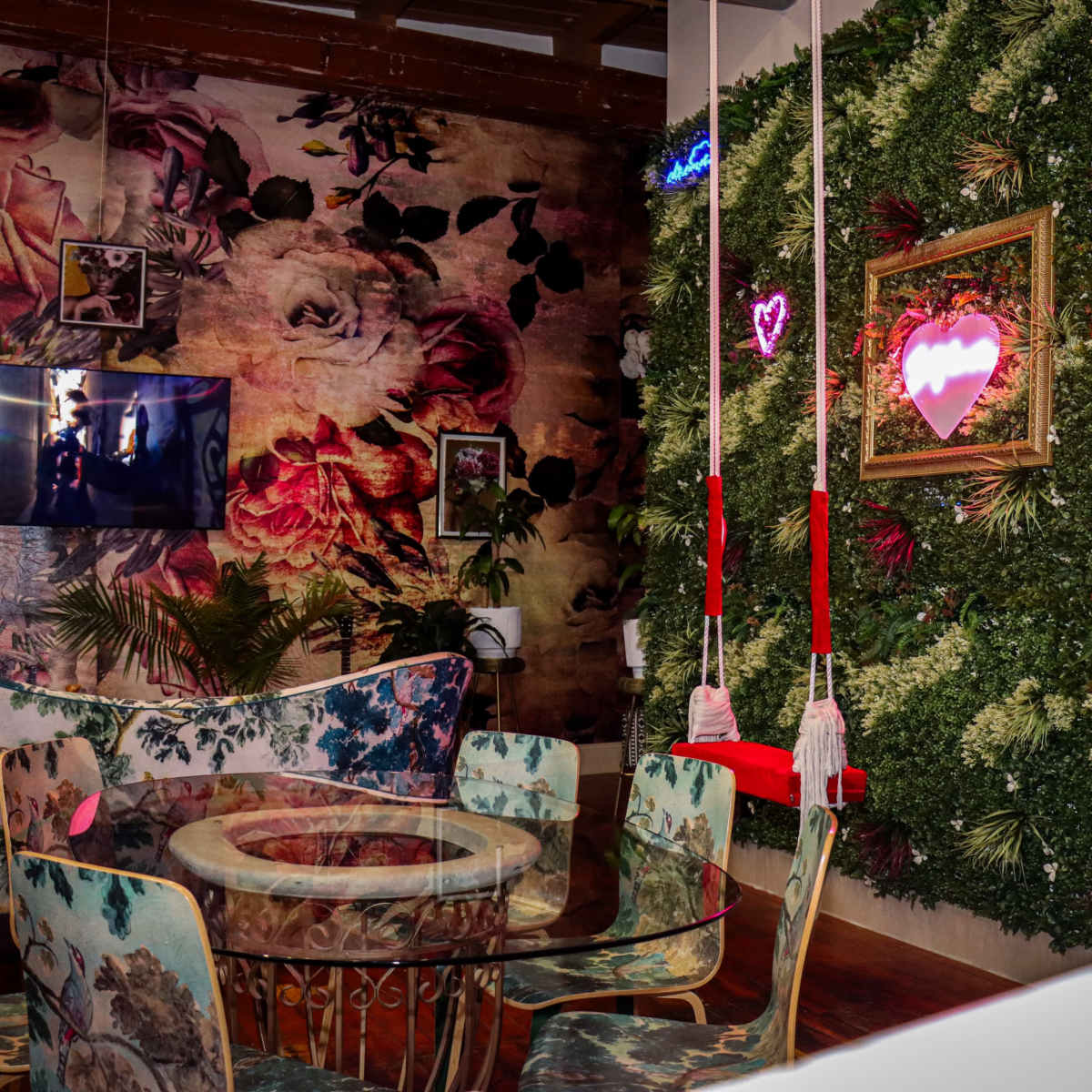 A vibrant interior of a café featuring floral wallpaper, a glass table, and a green ivy wall with neon heart decorations.