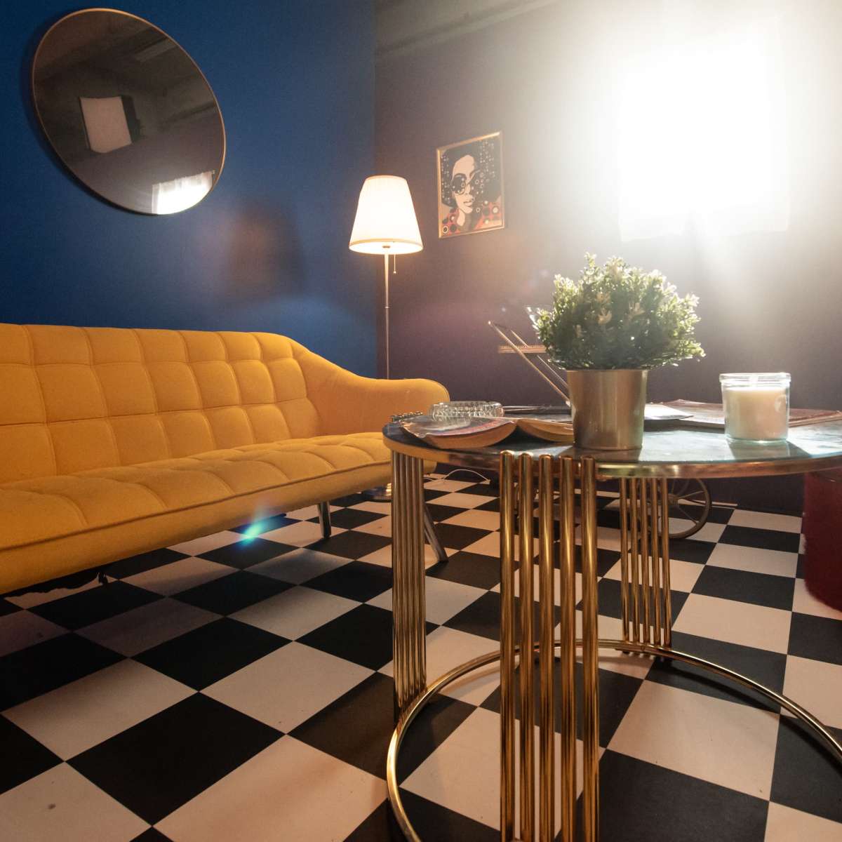A brightly lit room features a yellow sofa, a circular mirror, a coffee table with a plant and candle, and a checkered floor.