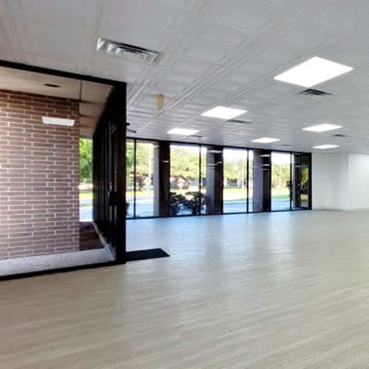 A spacious, empty room with white walls and a light wood floor, featuring large windows on one side.