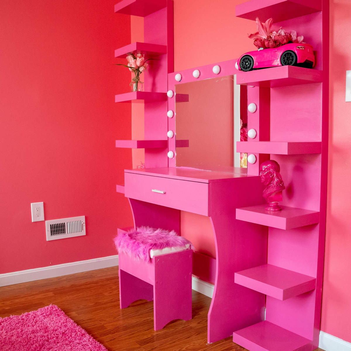A brightly colored pink vanity with shelves and a mirror is positioned against a pink wall in a room featuring a matching rug.