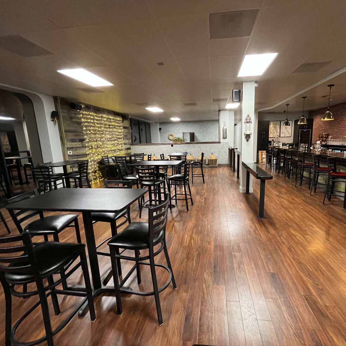 A spacious restaurant interior with wooden flooring, tables, and chairs arranged in a modern layout, featuring a bar area in the background.