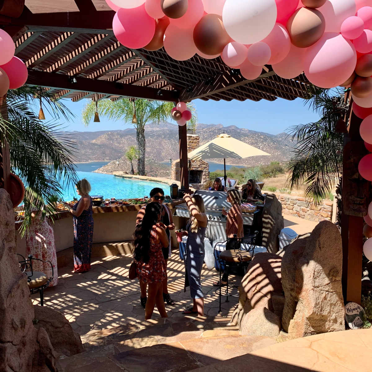 A group of people gather under a canopy adorned with pink and beige balloons, overlooking a pool surrounded by palm trees and hills.