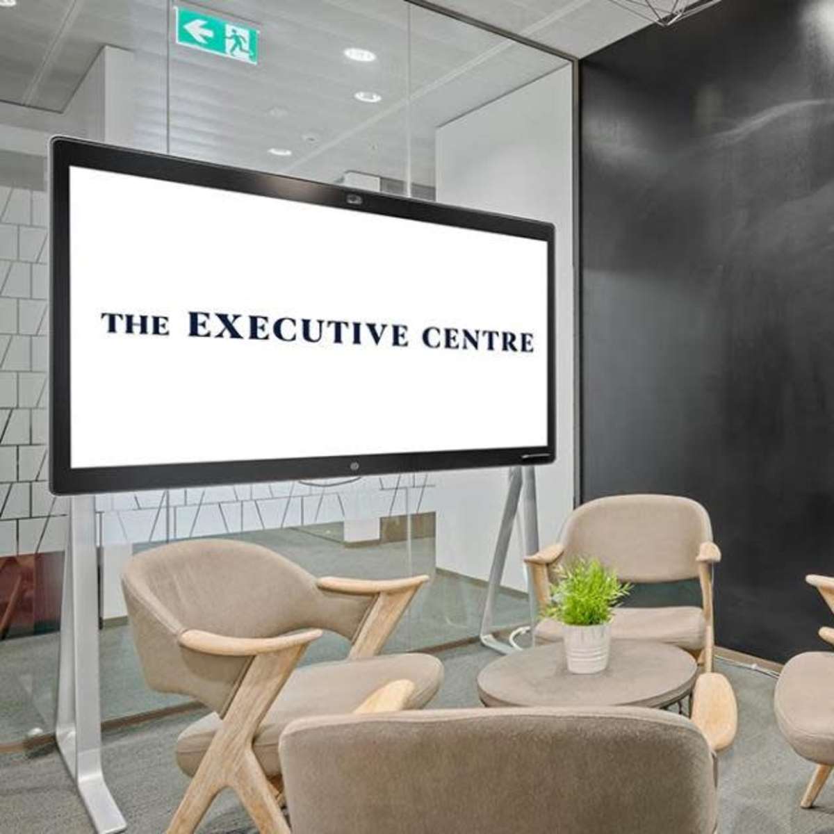 A modern meeting room features a large screen displaying "THE EXECUTIVE CENTRE" and includes four wooden chairs arranged around a small table.