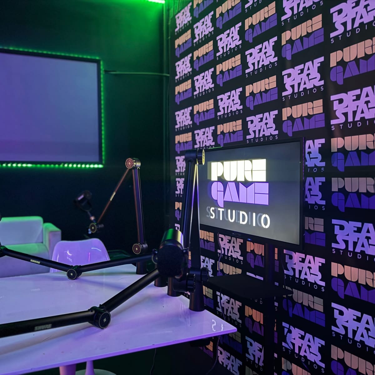 A modern podcast studio featuring a table with adjustable microphone arms and a wall branded with the name "PURE GAME STUDIO" illuminated by colorful LED lights.