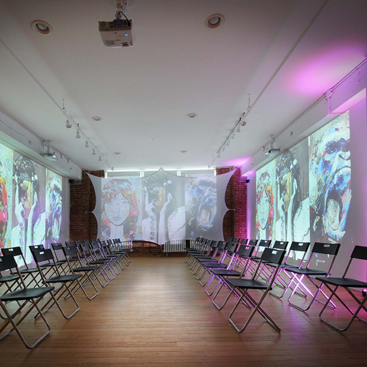 A spacious room with rows of black folding chairs facing multiple large projections on the walls, displaying colorful artistic images.