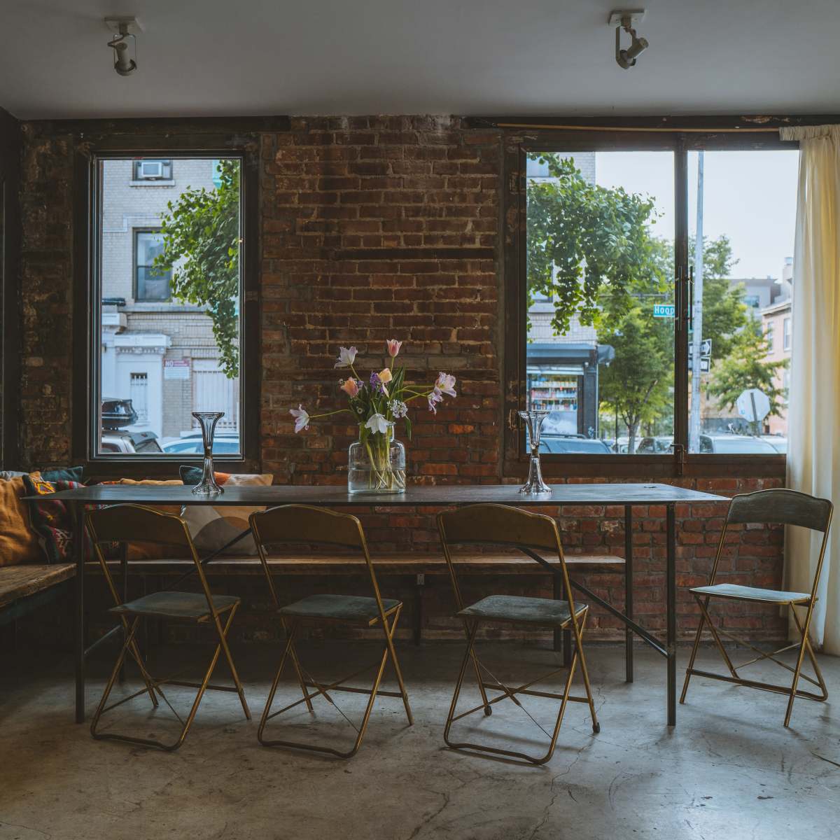 A long table with gold chairs sits in a room featuring exposed brick walls and large windows that let in natural light, complemented by a vase of flowers at the center.